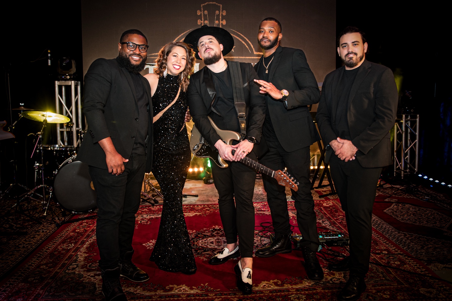 Tori and Will with their full band on stage, dressed in black with instruments and warm stage lights